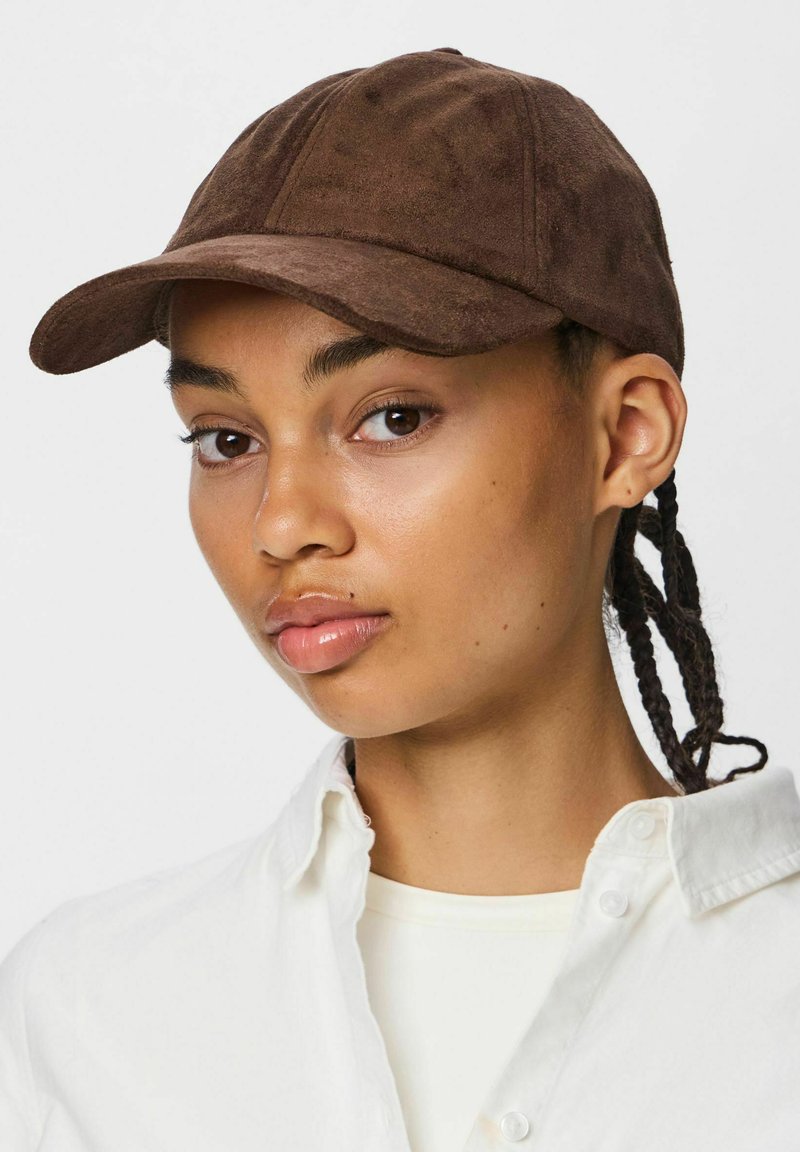 Jeune personne aux cheveux tressés portant une casquette de baseball en daim marron et une chemise blanche boutonnée sur un fond uni.