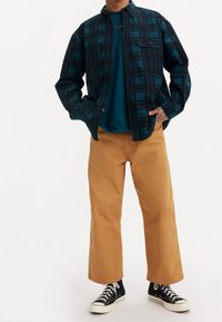 Kostkovaná košile v tmavě modrozelené a černé barvě přes modrozelené tričko. Neformální béžové kalhoty s rovnými nohavicemi a černé vysoké tenisky.