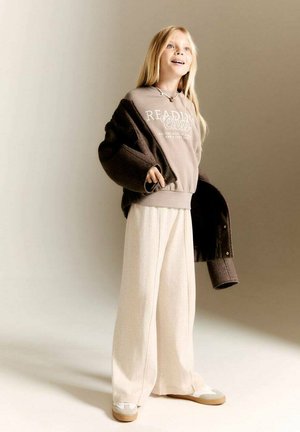 Sweatshirt marron clair avec le texte "Club de Lecture" superposé sous un manteau oversized marron foncé ; pantalon large crème ; baskets grises et blanches.