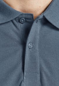 Šedé polo tričko s texturovanou tkaninou, klasickým límečkem a dvěma tmavými knoflíky na léze, které představuje úhledný a neformální design.