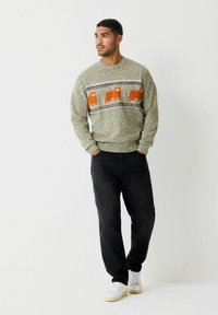 Zelený pletený sveter s oranžovými vzormi zvierat a prúžkom cez hruď, skombinovaný s tmavými džínsami a bielymi teniskami.