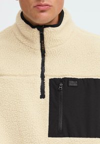 Puyo de forro polar beige con media cremallera y un bolsillo negro en el pecho, que presenta una cremallera suave y texturas contrastantes. Etiqueta del logo en el cuello.