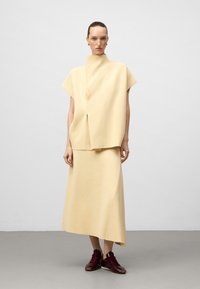 Femme portant un haut sans manches crème à col haut et une jupe asymétrique assortie, avec des baskets bordeaux, se tenant sur un sol gris.