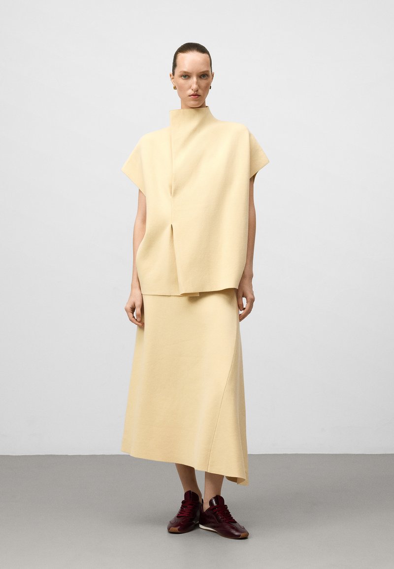Femme portant un haut sans manches crème à col haut et une jupe asymétrique assortie, avec des baskets bordeaux, se tenant sur un sol gris.