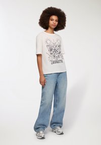 Biała graficzna koszulka z nadrukiem motyla i słońca, zestawiona z jasnoniebieskimi spodniami w szerokim kroju oraz srebrnymi trampkami. Prosta, teksturowana tkanina.