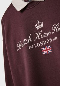 Bordo pulover s bež ovratnikom, ki vsebuje vezeno besedilo "Britanski konj R", krono in majhen oznako britanske zastave.