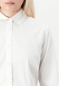 Woman wearing a plain white buttoned shirt with a classic collar, shown from neck to mid-torso against a white background.