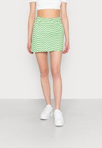 Saia mini verde com padrão, design em ondas brancas, material de denim, estilo de cinco bolsos e passadores de cinto. Combinada com ténis brancos.