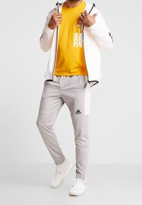 Weiße Zip-Jacke, gelbes T-Shirt mit Logo, graue Sweatpants mit schwarzen Akzenten und weiße Sneakers. Lässiges Sportoutfit.