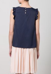 Navy blue sleeveless top with frill accents on shoulders, featuring three buttons at the back, paired with a pleated light pink skirt.