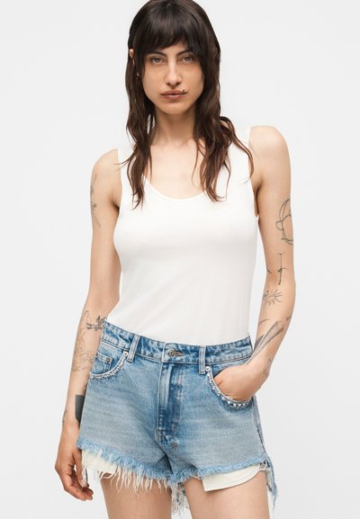 Γυναίκα με σκούρα μαλλιά που φοράει λευκή μπλούζα χωρίς μανίκια και ξεθωριασμένο μπλε τζιν σορτς με τσέπες με τρουκς, στέκεται με το ένα χέρι στην τσέπη.