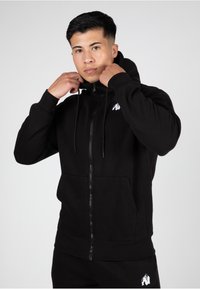 Zwarte zip-up hoodie van zachte stof, met een voorkantzak, trekkoorden en een klein wit logo op de linkerkant van de borst.