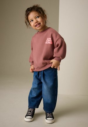 Lyserød sweatshirt med teksten "Let Love Bloom", kombineret med løse blå denimjeans og sorte sneakers med hvide såler og et blomsterprint.