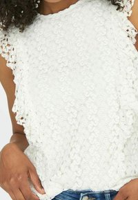 Weißes Spitzenoberteil mit floralen Mustern, strukturiertem Stoff und gewellten Kanten, kombiniert mit Denim-Shorts.