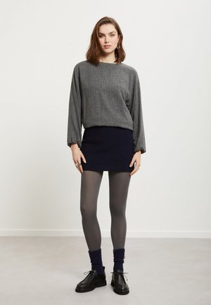 Mujer de pie, vistiendo un jersey gris de manga larga, falda mini negra, medias grises, calcetines azul marino y zapatos negros con cordones, sobre un fondo blanco liso.