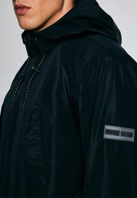 Tuva cilvēka melnas kapuces jaku ar rāvējslēdzēju un regulējamām auklām, uz piedurknes ar taisnstūra formu "BIG STAR" uzlīmi.