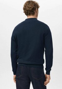 Granatowy dziergany sweter z długimi rękawami, ściągaczowymi mankietami i okrągłym dekoltem. Teksturowana tkanina, swobodny krój. Noszony z ciemnymi dżinsami.