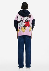 Rosa Strick-Hoodie mit Disney-Charakteren Micky und Minnie Maus, mit dunkelblauen Ärmeln und einem lässigen Schnitt, kombiniert mit dunkelblauen Jeans.