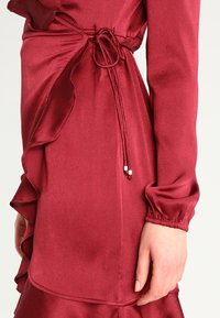 Robe bordeaux avec ourlet et manches volants. Texture satinée, détail de taille noué avec deux accents argentés. Design lisse et ajusté.