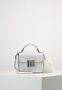 Sac à main argenté scintillant avec un fermoir métallique, des accents noirs et un charm en fausse fourrure blanche amovible. Comprend une poignée supérieure et une bandoulière ajustable.