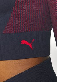 Marineblaues Sportoberteil mit strukturierten roten Mustern und gestricktem Design. Ein rotes Puma-Logo ist entlang dem Saum sichtbar. Glattes Material mit taillierter Silhouette.
