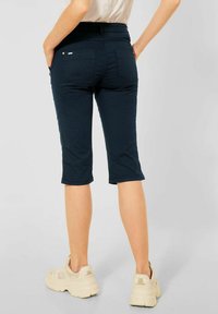 Mörkblå capribyxor med en figurnära design, två bakfickor och subtila sömdetaljer, tillsammans med ljusa sneakers.