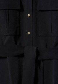 Vêtement noir avec ceinture en tissu nouée à la taille, deux poches avant à rabat et deux boutons-pression métalliques visibles au centre.