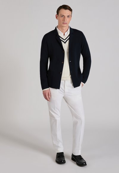 Navy knit jacket with buttons, layered over a cream cable-knit sweater, paired with white trousers and black shoes against a neutral background.