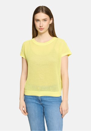 Jonge vrouw met steil bruin haar die een geel gebreid topje met korte mouwen en een blauwe spijkerbroek draagt, staande tegen een effen achtergrond.