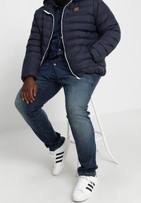 Man die op een witte kruk zit, gekleed in een marineblauwe pufferjack, een donkerblauwe overhemd, blauwe jeans en witte sneakers met zwarte strepen.