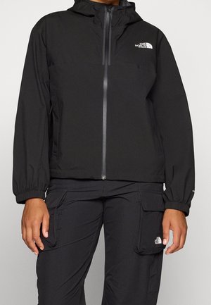 Osoba ubrana w czarną kurtkę z kapturem zapinaną na zamek błyskawiczny oraz czarne spodnie cargo, obie z logo The North Face na piersi i kieszeni na udzie.