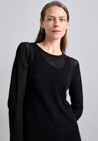 Maglietta nera a maniche lunghe in tessuto mesh, con scollatura a girocollo e motivo testurizzato. Il design è aderente con una silhouette slanciata.