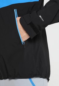 Main dans la poche d'une veste noire et bleue montrant l'étiquette DRYVENT sur la manche avec une sangle Velcro ajustable.