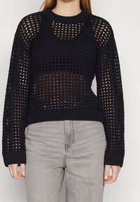 Maglione nero a maglia con ampio design a rete, colletto rotondo, maniche lunghe e orlo a costine. La texture è a maglia aperta, rivelando la pelle sottostante.