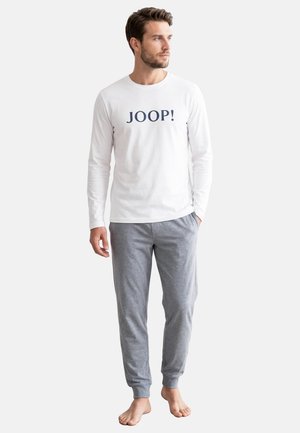 Mężczyzna w białej koszuli z długim rękawem z napisem „JOOP!” i szarych spodniach dresowych, stojący boso na jednolitym tle.