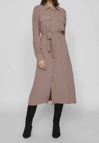 Dlouhý rukáv, košilové šaty v měkké mauve látce, s dvěma kapsami na prsou, páskem v oblasti pasu a rovnými střihy, kombinované s černými kozačkami ke kolenům.