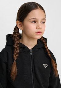 Czarna bluza z kapturem na zamek z gładką fakturą i małym haftowanym wzorem na piersi, z kapturem i fryzurą w warkocz boczny.