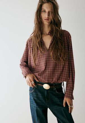 Femme aux cheveux longs portant une chemise à carreaux rouge rentrée dans un jean bleu avec une grande boucle de ceinture ovale et des bagues aux doigts.