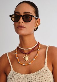 Γυναίκα που φοράει μαύρα γυαλιά ηλίου, πολλά χρυσά σκουλαρίκια και πολλάπλές κολιέ, συμπεριλαμβανομένου ενός χρυσού μενταγιόν χελώνας, και ένα μπεζ πλεκτό τοπ.