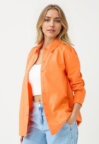 Camicia arancione con colletto aperto, maniche lunghe e una texture liscia, indossata sopra un crop top bianco e abbinata a jeans chiari.