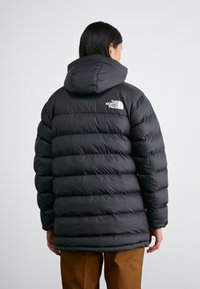 Giacca a vento nera con sezioni trapuntate orizzontali e cappuccio. Presenta un logo bianco sulla parte superiore della schiena. Materiale caldo e design casual.