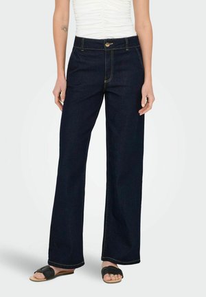 Jean large en denim foncé avec des coutures jaunes et une fermeture à bouton à l'avant, assorti à un haut blanc ajusté et des sandales noires.
