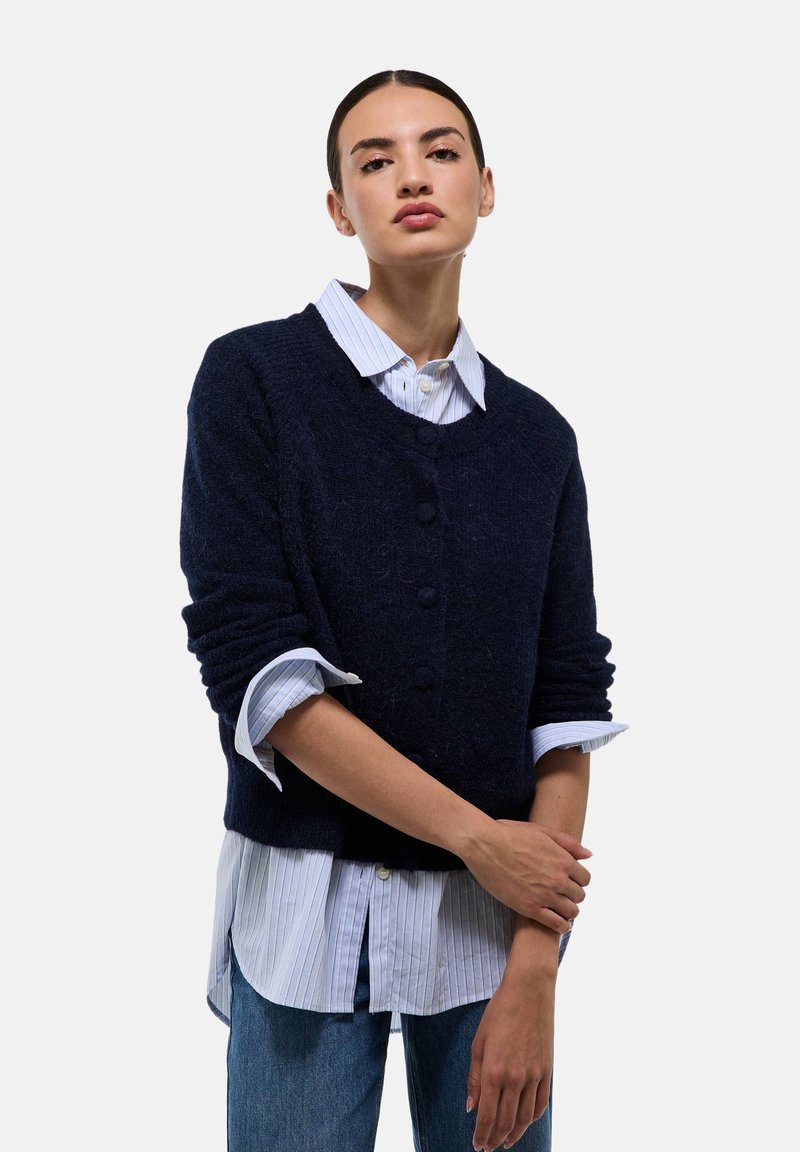 Pull en maille bleu marine texturé avec un col en V et fermeture à boutons, superposé à une chemise blanche à rayures. Coupe décontractée avec des manches retroussées.