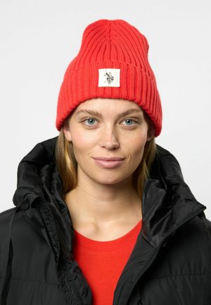 Raudona ripsuota megzta kepurė su siuvinėta balta etikete, kurioje pavaizduotas grafikos logotipas; dėvima su juoda su gobtuvu striuke virš raudonos marškinėlių.