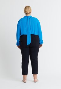 Frau mit blondem Haar in einem niedrigen Dutt trägt eine leuchtend blaue Bluse mit Bindeband-Detail, schwarze, gekürzte Hosen und silberne Sandalen, steht mit dem Rücken zur Kamera.