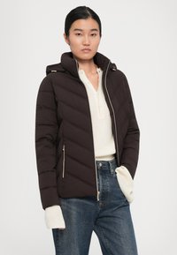 En ung kvinne iført en mørkebrun quiltet jakke over en kremfarget strikkegenser og blå jeans, står foran en enkel hvit bakgrunn.
