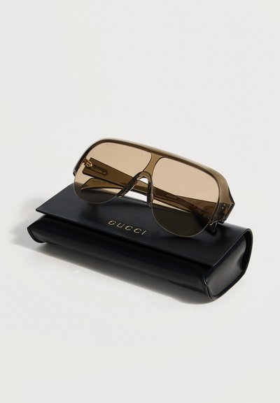Καφέ αποχρωματισμένα υπερμεγέθη γυαλιά ηλίου με διαφανή σκελετό που ακουμπούν σε μαύρη θήκη Gucci με χρυσά γράμματα λογότυπου.