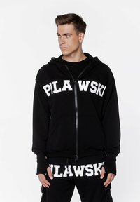 Czarna bluza z kapturem zapinana na zamek, wykonana z mi ękkiego materiału, z dużym białym napisem "PLAWSKI" na klatce piersiowej. Model ma na sobie dopasowane spodnie.