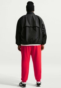 Zwarte nylon jas met een losse pasvorm, elastische manchetten en een grote achterklep; gecombineerd met rode joggingbroek en zwarte sneakers met witte accenten.