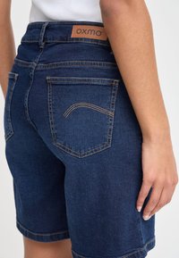 Tamsiai mėlynos spalvos džinsiniai šortai su vidutinio aukščio liemeniu, užpakalinėmis kišenėmis ir kontrastingomis siūlėmis. Odinis prekės ženklo ženklas matomas nugaroje.